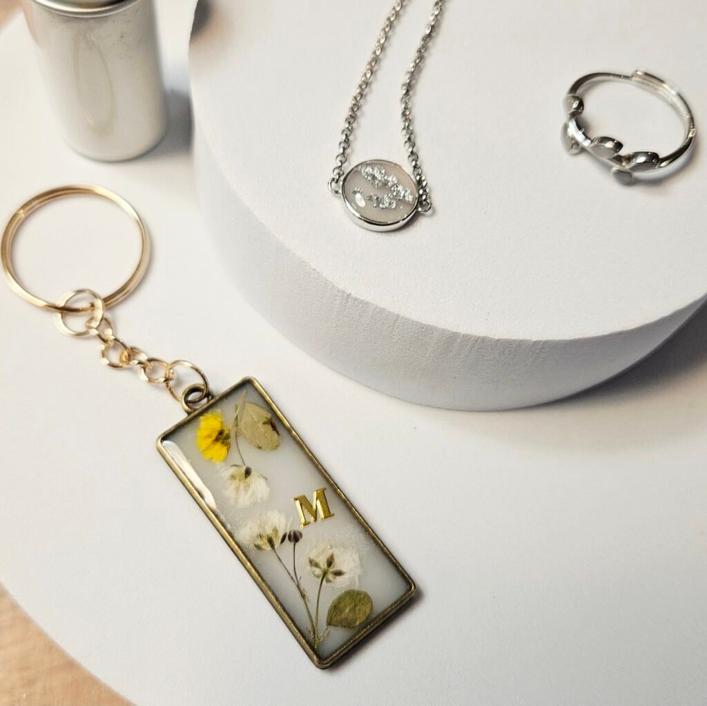 Muttermilchschmuck: Schlüsselanhänger mit getrockneten Blüten und goldenem Initial „M", dazu Kette und Ring in Silber, aus dem Workshop in Wuppertal