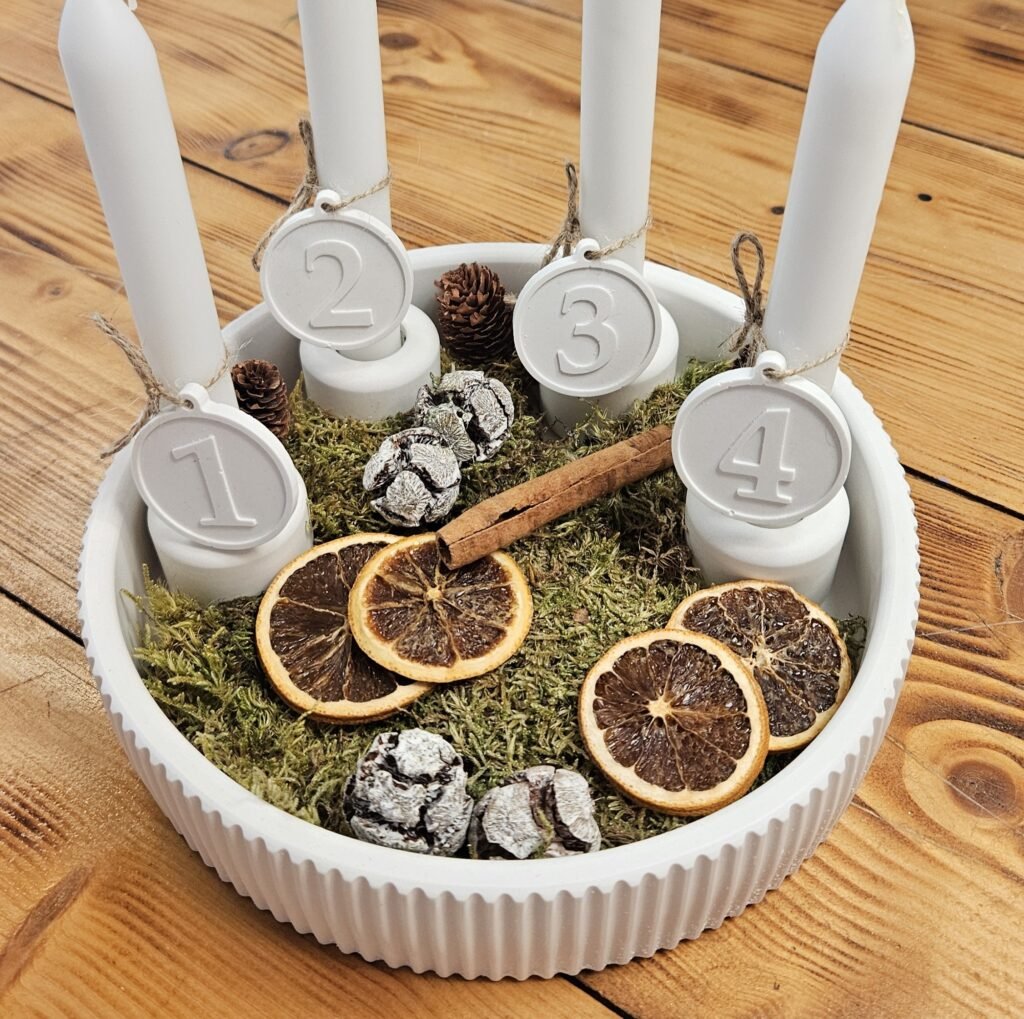 Adventskranz aus Gießkeramik: gerillte weiße Schale mit vier Kerzen, Zahlenanhängern 1–4, Moos, Orangenscheiben und Zapfen