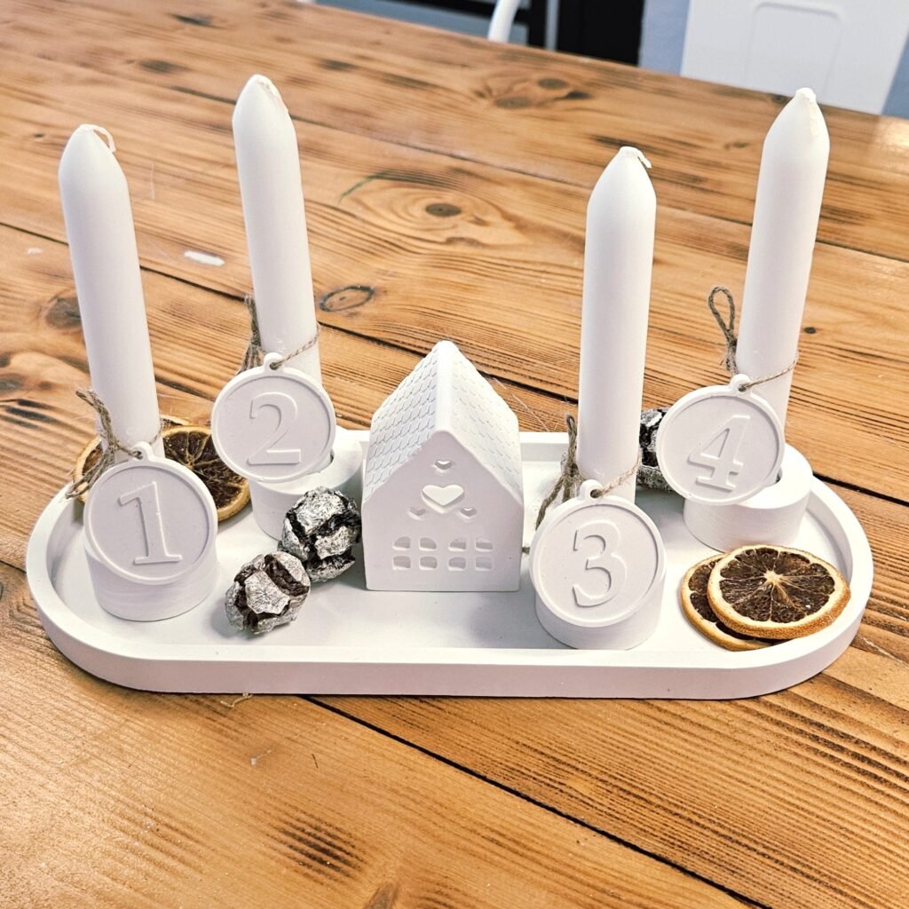 Weißes Adventskranz-Tablett aus Gießkeramik mit Keramikhaus, Kerzen, Zahlenanhängern an Jutekordel und getrockneten Orangen