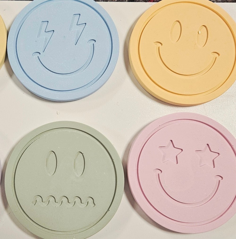 Vier runde Untersetzer aus Gießkeramik mit Smiley-Relief in Hellblau, Gelb, Salbeigrün und Rosa, aus dem Workshop Gießkeramik in Wuppertal