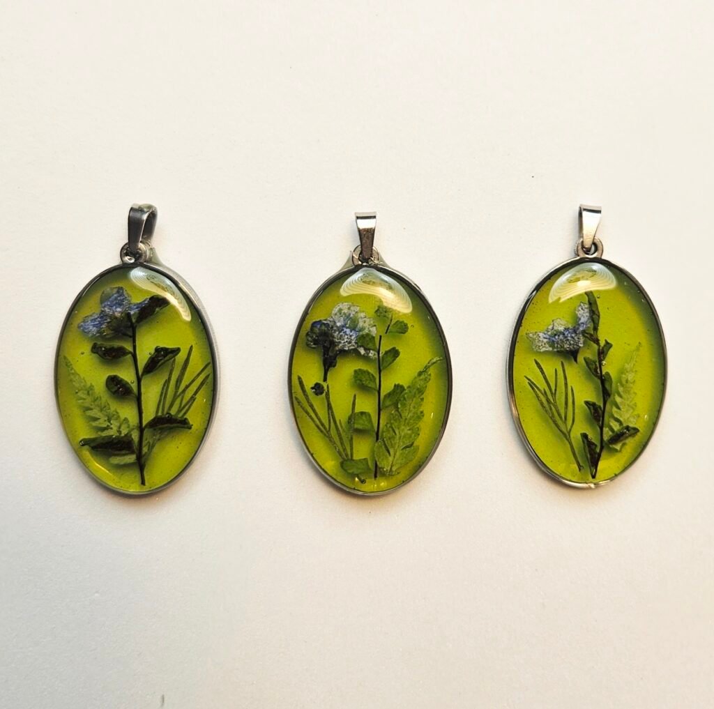 Drei ovale UV-Resin Anhänger mit gepressten Blüten und Farnblättern auf hellgrünem Grund in Silberfassung, aus dem Schmuck-Workshop in Wuppertal
