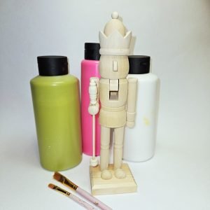 Unbemalter Nussknacker aus hellem Holz mit Krone und Zepter vor Acrylfarbflaschen in Olivgrün, Pink und Weiß, daneben zwei Pinsel